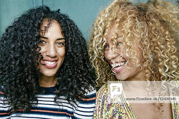 Porträt von zwei jungen Frauen mit langen lockigen schwarzen und blonden Haaren  die in die Kamera lächeln.