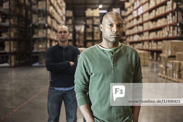 Portrait of an African American and Caucasian warehouse workers in a large distribution warehouse with racks of products stored in cardboard boxes on racks in the background.