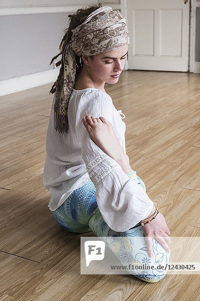 Junge Frau mit Kopftuch und weißer Bluse sitzt in Yoga-Pose auf dem Boden.