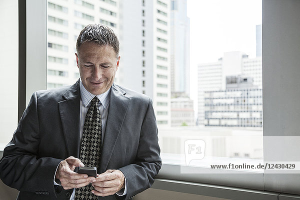 A Caucasian businessman texting on his phone while standing next to a window.