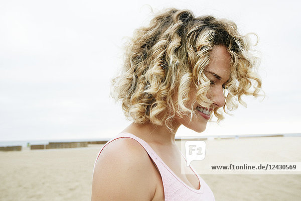 Porträt einer lächelnden jungen Frau mit blonden Locken  die am Sandstrand steht.