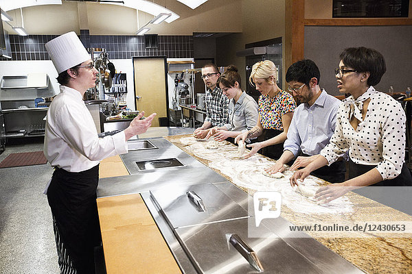 A Caucasian male chef teaching a cooking class for a mixed race grope of students in a commercial kitchen