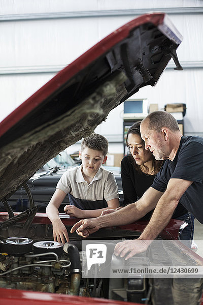 A senior Caucasian male car mechanic talks to a Caucasian mother and her son about a engine repair issue in a classic car repair shop.