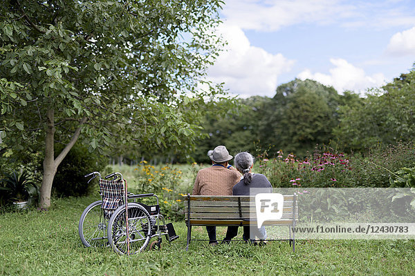 Ehemann und Ehefrau  Rückansicht eines älteren Mannes mit Hut und einer Frau  die Seite an Seite auf einer Bank in einem Garten sitzen  neben der ein Rollstuhl geparkt ist.