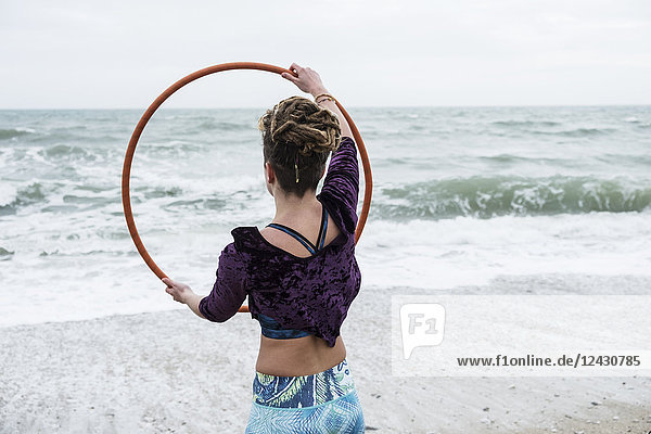 Junge Frau mit braunen Haaren und Dreadlocks  die auf einem Sandstrand am Meer steht und Hula-Hoop-Reifen balanciert.
