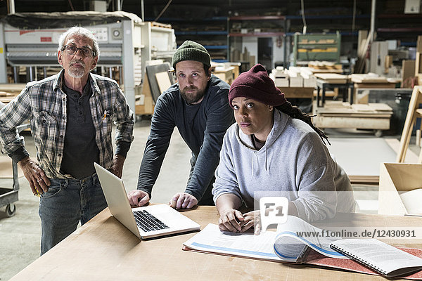 A portrait of mixed race carpenters around a laptop computer after work hours in a large woodworking facility.