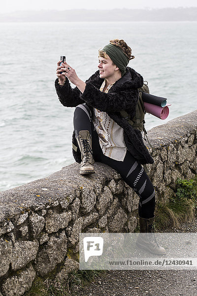 Young woman with brown hair and dreadlocks wearing headscarf sitting on dry-stone wall near ocean  taking picture with her mobile phone.