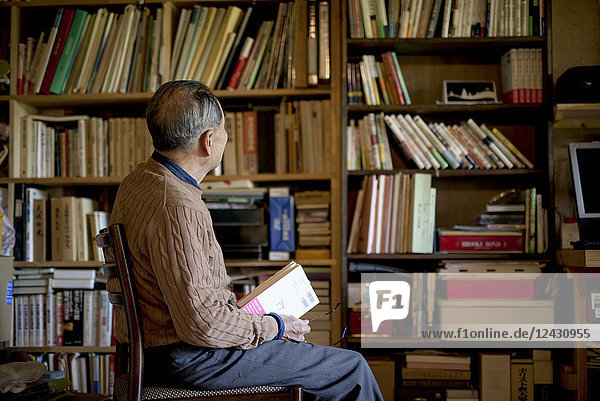 Älterer Mann sitzt auf einem Stuhl vor einem Bücherregal und hält ein Buch.