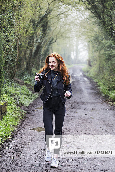 Lächelnde junge Frau mit langen roten Haaren  die einen Waldweg entlang läuft und mit einer Oldtimer-Kamera fotografiert.