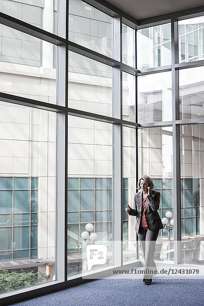 A Caucasian businesswoman talking on a cell phone while standing next to a large window in a convention centre lobby.