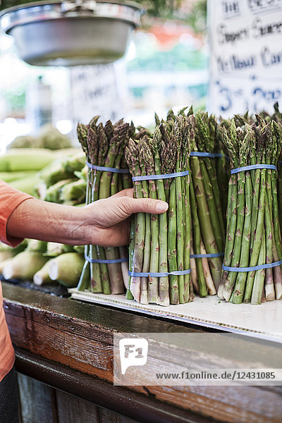 Nahaufnahme einer menschlichen Hand  die auf einem Obst- und Gemüsemarkt einen Bund frischen grünen Spargels hält.
