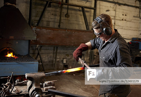 Artisan metal worker wearing ear protectors and goggles using a hammer and anvil to shape a red hot piece of metal with a twist in the piece.