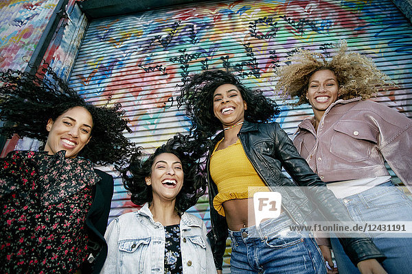 Niedrigwinkelaufnahme von vier jungen Frauen mit lockigem Haar  die mit bunten Graffiti bedeckt vor dem Fensterladen stehen und in die Kamera lächeln.