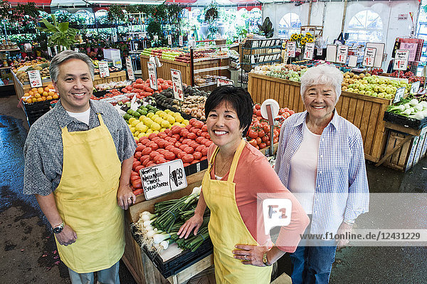 Schrägaufnahme eines Mannes und zweier Frauen mit Schürzen  die an einem Stand mit frischen Tomaten auf einem Obst- und Gemüsemarkt stehen.