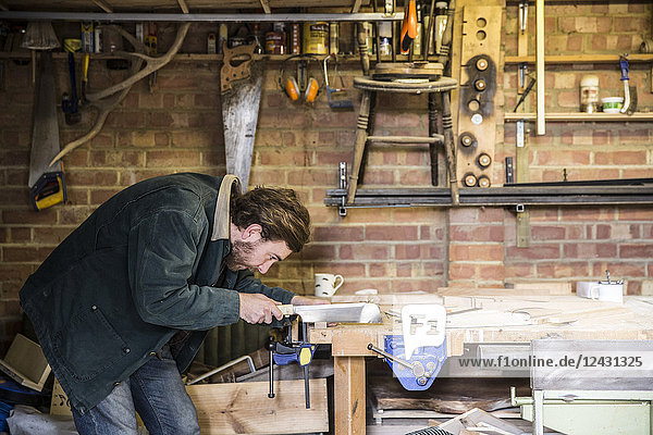 Mann arbeitet stehend an einer Werkbank in einer Holzbearbeitungswerkstatt  mit einer Säge auf einem Stück Holz.