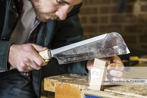Nahaufnahme eines Mannes  der stehend in einer Holzwerkstatt arbeitet  mit einer Säge auf einem Stück Holz.