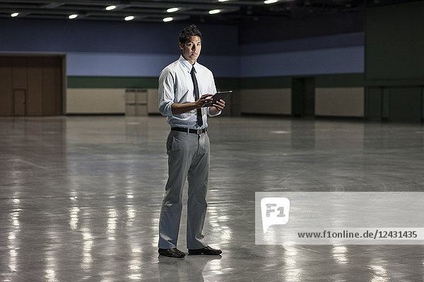 Portrait of a businessman holding a notebook computer and standing in a dimly lit and dark exhibition area in a convention centre.
