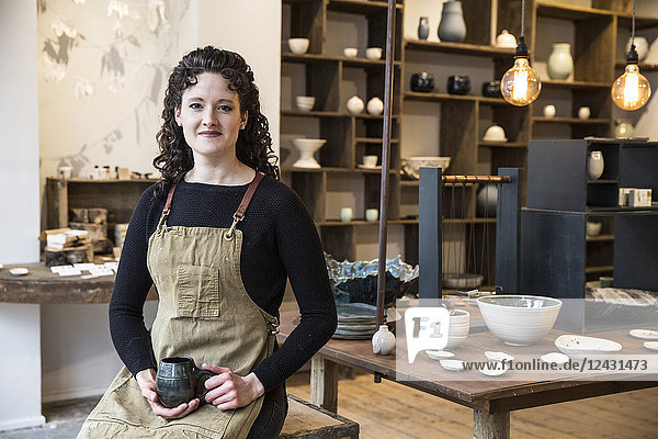 Woman with curly brown hair wearing apron standing in her pottery shop  holding mug  smiling at camera.