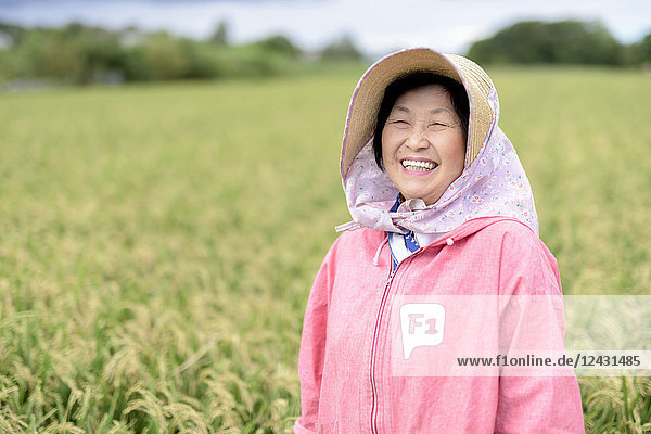 Lächelnde Frau mit Strohhut und rosa Jacke steht in einem Reisfeld und schaut in die Kamera.