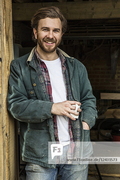 Bearded man standing in doorway of woodworking workshop  holding mug  smiling at camera.