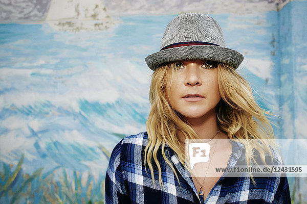Portrait of woman with long blond hair wearing blue checked shirt and grey Trilby hat  looking at camera.