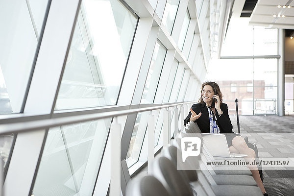 Caucasian businesswoman using her cell phone while sitting in a row of chairs against a window in a conference centre lobby.