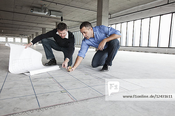 Ein Architekt und ein Geschäftsinhaber kauern über Architekturplänen und zeichnen auf einen Betonboden und planen einen neuen Büroraum in einem leeren  roh gebauten Raum.