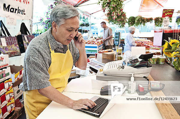 Mann mit Schürze steht an der Kasse eines Lebensmittel- und Gemüsemarktes  telefoniert  tippt am Laptop.
