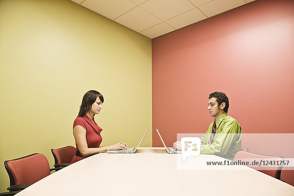 Ein spanischer Mann und eine kaukasische Frau auf Schoßcomputern in einem farbenfrohen Bürokonferenzraum.