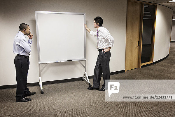A black businessman and an Asian businessman ponder the issue of an empty white board.