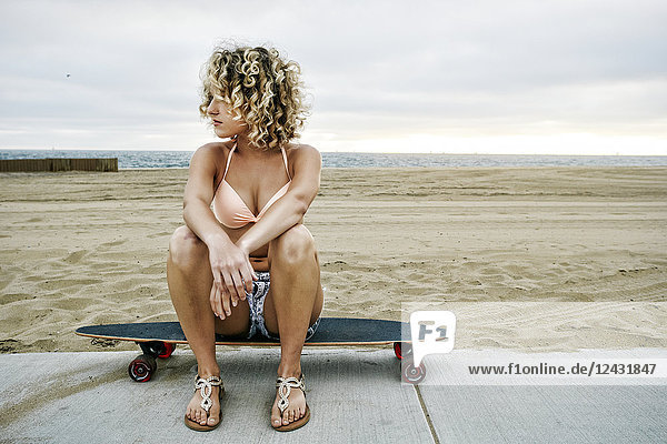 Junge Frau mit lockigem blonden Haar im rosa Bikini  die auf einem Skateboard am Sandstrand sitzt.