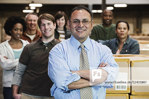 Team portrait of multi-ethnic warehouse workers lead by a Hispanic American male executive and surrounded by large racks of products stored in cardboard boxes in a large distribution warehouse.