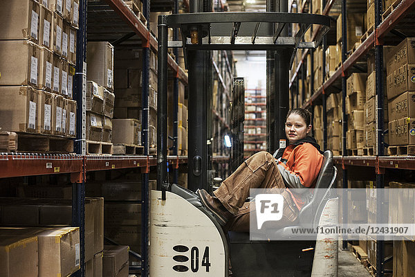 Caucasian female warehouse worker sitting in a motorized stock picker surrounded by products stored in cardboard boxes in a large warehouse distribution facility.