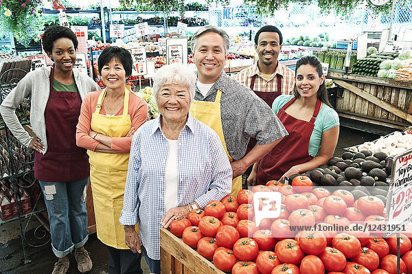 Schrägaufnahme einer Gruppe von Männern und Frauen mit Schürzen  die an einem Stand mit frischen Tomaten auf einem Obst- und Gemüsemarkt stehen.