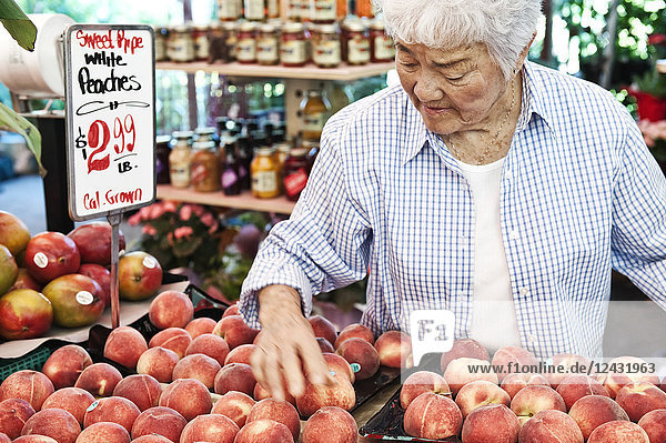 Ältere Frau steht an einem Stand mit frischen weißen Pfirsichen auf einem Obst- und Gemüsemarkt.