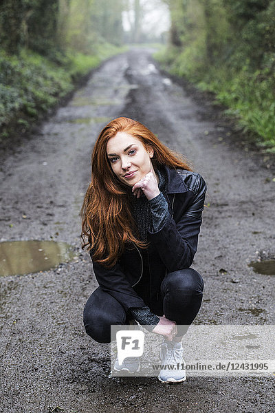 Porträt einer lächelnden jungen Frau mit langen roten Haaren  die auf einem Waldweg kniet und in die Kamera schaut.