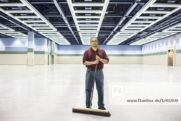 An Hispanic man standing with a broom in a large convention cener space.