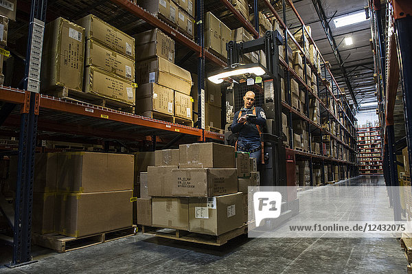 Warehouse worker wearing a safety harness while operating a motorized stock picker in an aisle between large racks of cardboard boxes holding product on pallets in a large distribution warehouse.