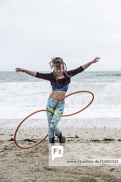 Young woman with brown hair and dreadlocks standing on a sandy beach by the ocean  balancing two hula hoops.