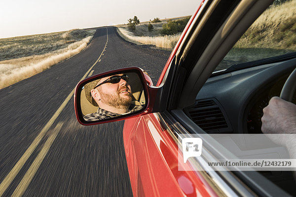 View in the rear view drivers mirror of a Caucasian male driving a car on a road trip in eastern Washington State  USA