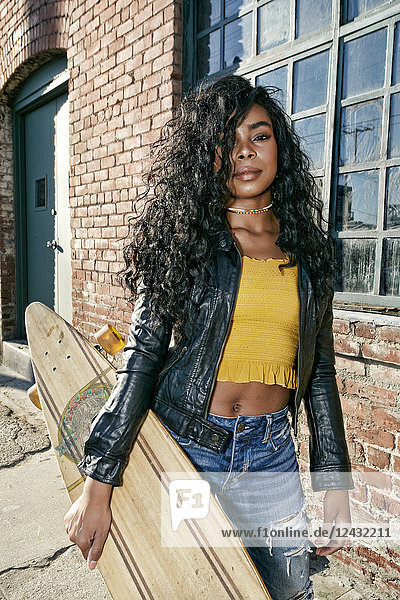 Young smiling woman with long curly black hair standing on pavement  holding skateboard  looking at camera.