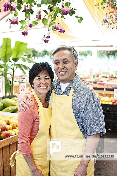 Ein Mann und eine Frau  die einen Obst- und Gemüsemarkt betreiben  tragen Schürzen  stehen nebeneinander und lächeln in die Kamera.