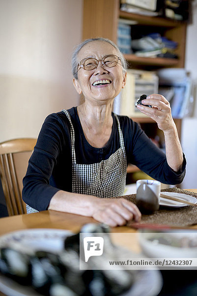 Ältere Frau sitzt am Küchentisch  isst Sushi und lächelt in die Kamera.
