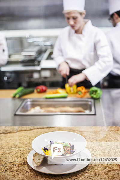 Eine Nahaufnahme eines fertigen Tellers mit Köchen  die im Hintergrund in einer Großküche arbeiten