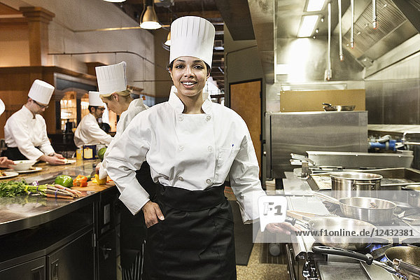 A portrait of a young black female chef in a commercial kitchen with her crew working behind her.