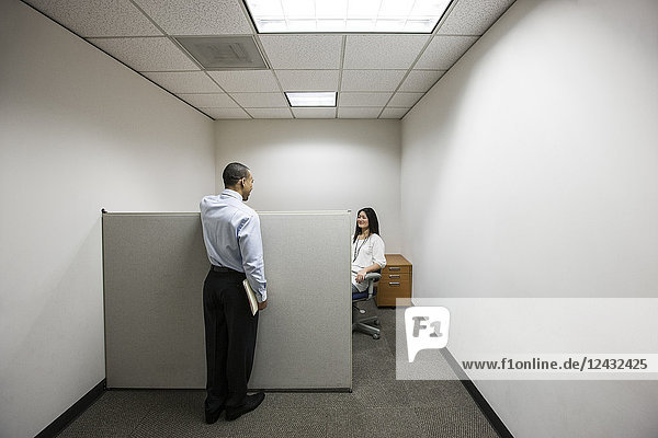 A black businessman talking to an Asian businesswoman working in a small corner cubicle office.