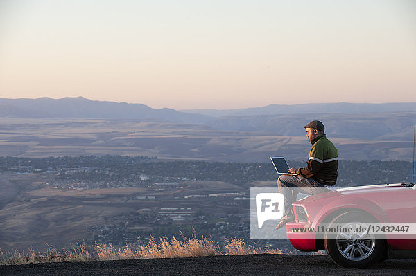 Kaukasischer Mann  der auf der Motorhaube seines Autos sitzt und an einem Laptop-Computer auf einem Rastplatz in der Nähe von Lewiston  Idaho  USA  arbeitet.