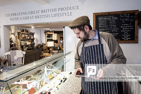 Bearded man wearing flat cap and apron standing at counter in a delicatessen  holding digital tablet.