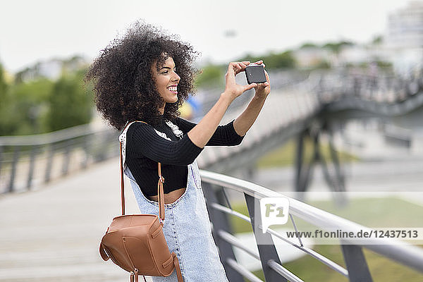 Smiling young woman with brown leather backpack standing on a bridge taking photos with smartphone