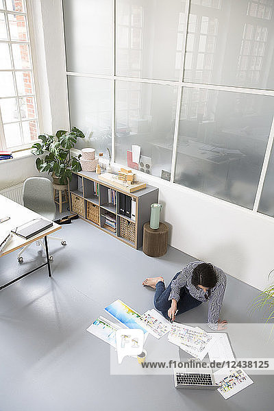 Casual woman with sheets of paper and laptop sitting on the floor in a loft office